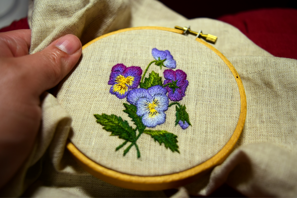 Hand embroidered design of blue and purple pansies, nearly finished in hoop