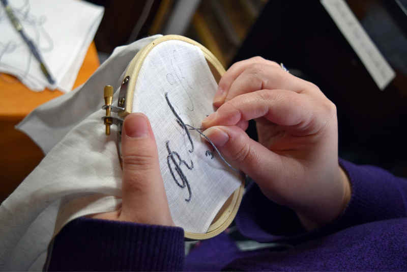 Embroidered letters in progress in hoop, a woman in a purple shirt is embroidering them by hand onto a linen handkerchief