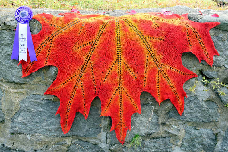 Hand spun, hand knit, and hand dyed shawl shaped to look like a scarlet maple leaf with gold veins, posed on rock wall with Best of Department ribbon from the Eastern States Expo alongside