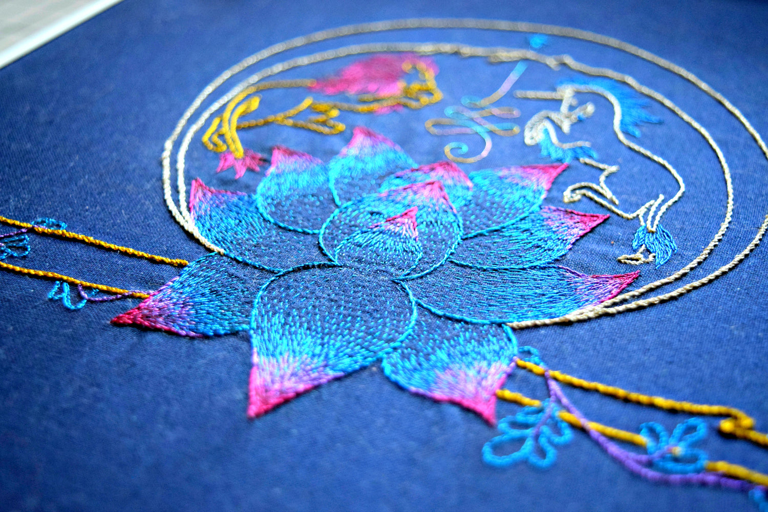 "Journey Talisman" hand embroidered design worked in fine metallic threads on blue background. A shimmering lotus sits between a lion and unicorn encased in a sphere and atop a golden bough