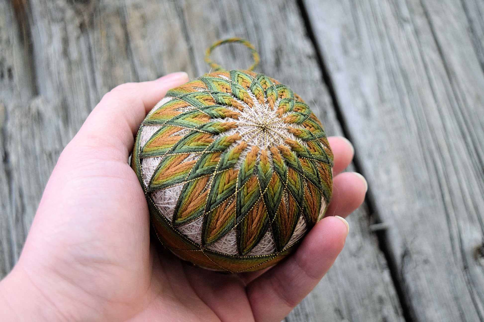 Woman's hand holding temari ball work in gold, green, and brown