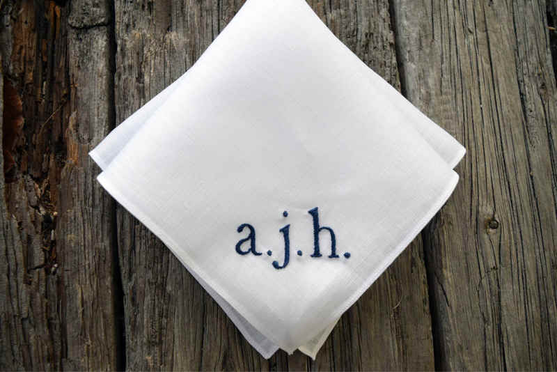 Folded white linen pocket square with hand embroidered navy initials (lowercase) a. j. h. 