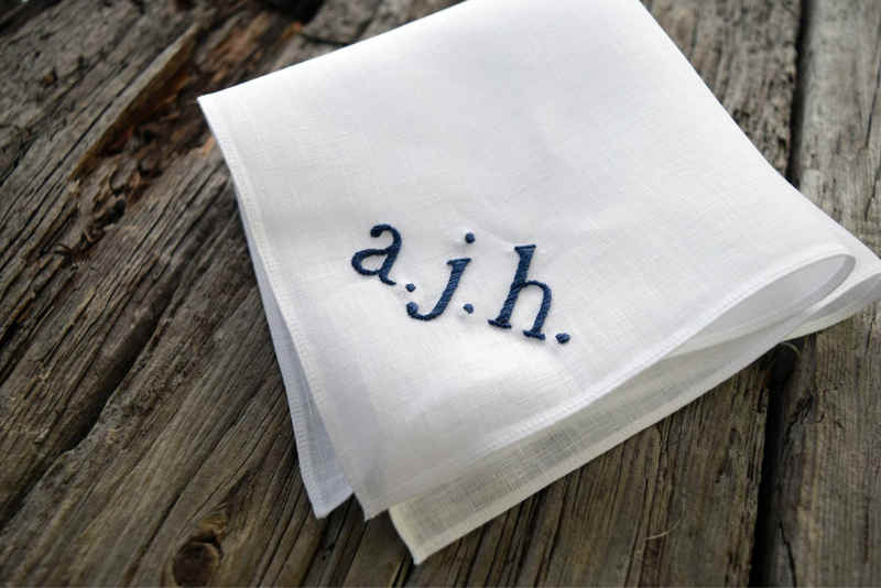 Side view of white Irish linen hankie embroidered with three lowercase navy initials in serif font. Initials read a. j. h.