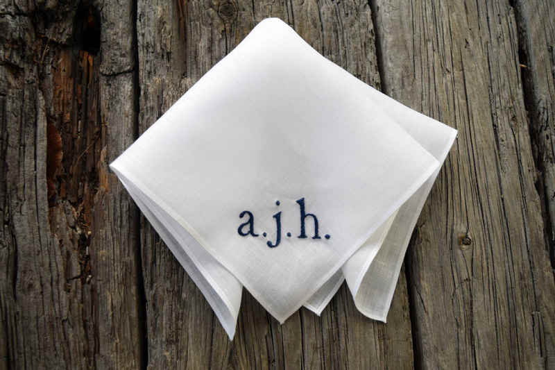 White Irish linen handkerchief on wood background hand embroidered with three navy initials, lowercase. Initials read a. j. h.