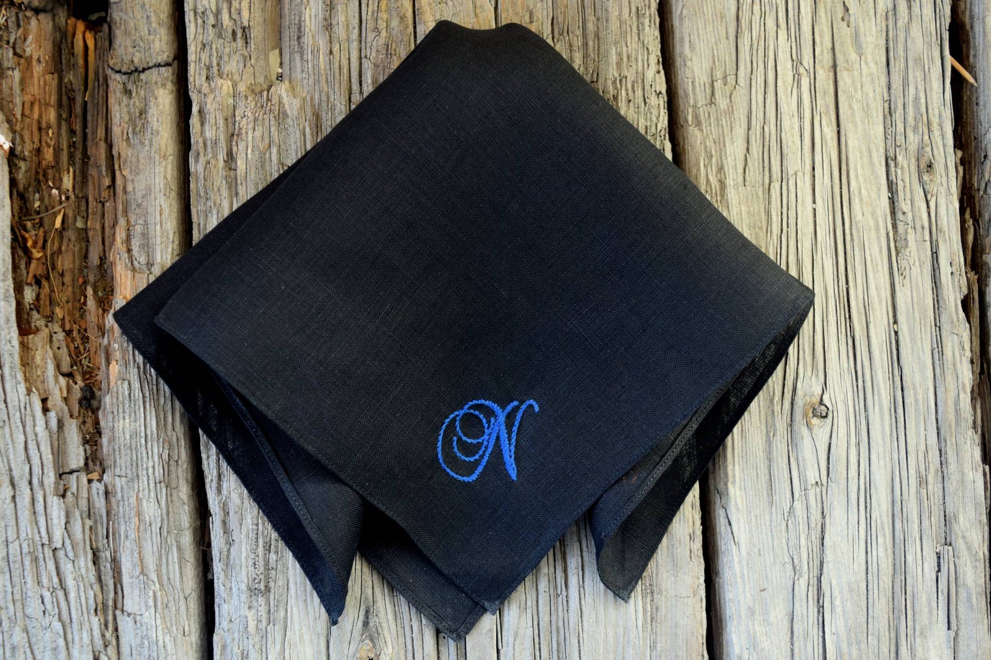 Black linen hankie on wood background hand embroidered with a letter N in blue