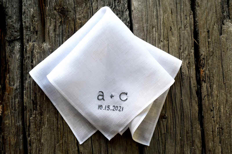 White Irish linen handkerchief casually folded on wood background. Handkerchief is embroidered with two letters and a wedding date, in grey, in chain stitch.