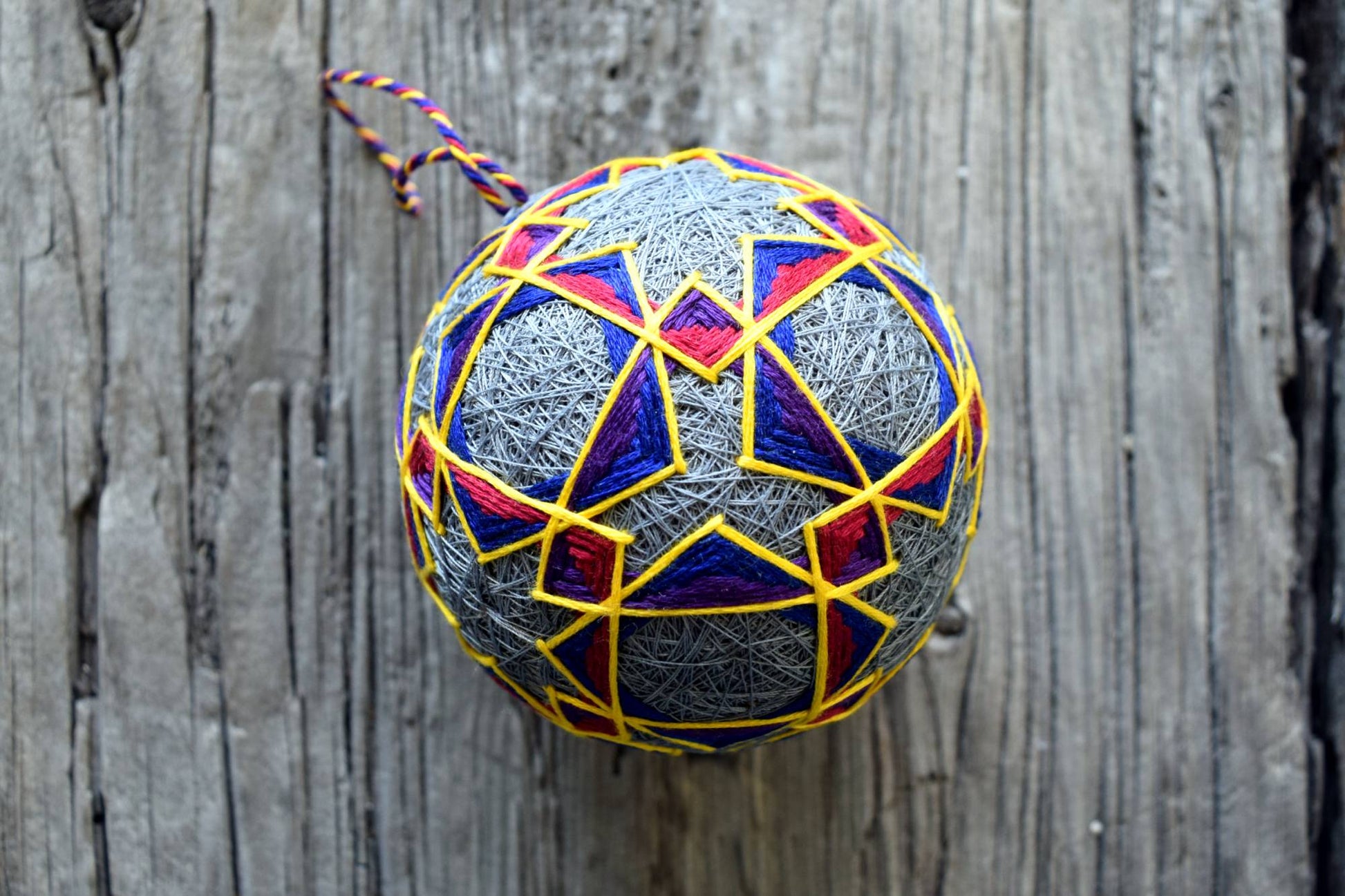 Closeup of Japanese temari ball in red, purple, blue, yellow on grey background. Design of interlocking shapes.