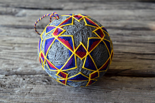 Closeup of Japanese temari ball with twisted thread loop. Base of design grey, embroidery in blue, purple, red, and golden yellow forming interlocking shapes