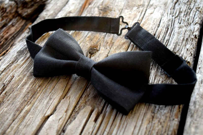 Side view of black linen bow tie lying on wood. Adjustable fastener at back of neck is visible.