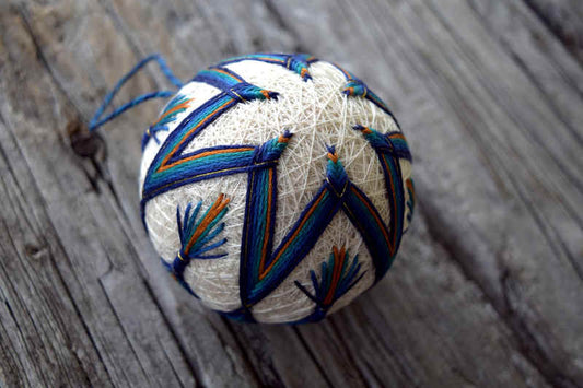 Cream temari embroidered in shades of blue, teal, and ochre in six pointed flower/star pattern. Fan shaped stitches around the points suggest peacock feather shapes.