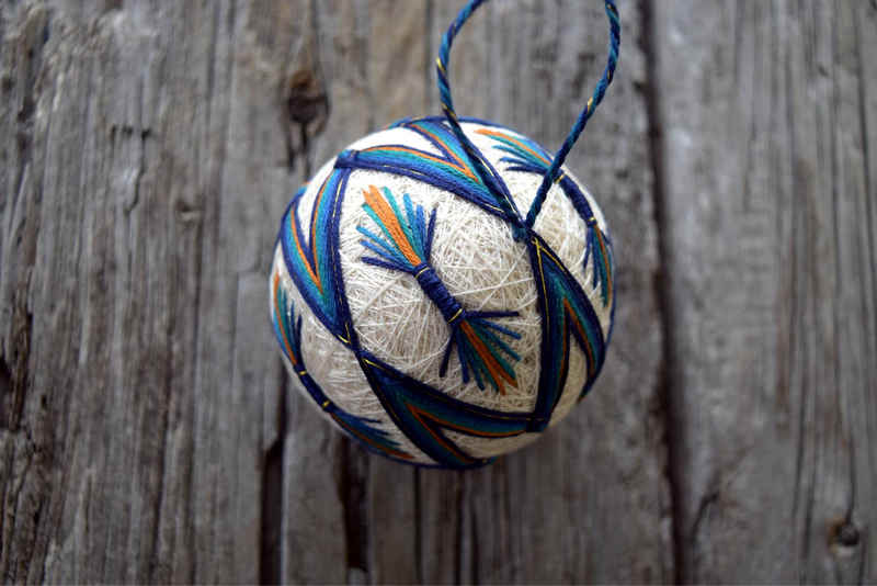 Temari ball 'Peacock' viewed from side near thread loop hanger; bundles of straight stitches are gathered with a deep blue and gold band so they resemble double-pointed feathers against the cream base of the temari ball.