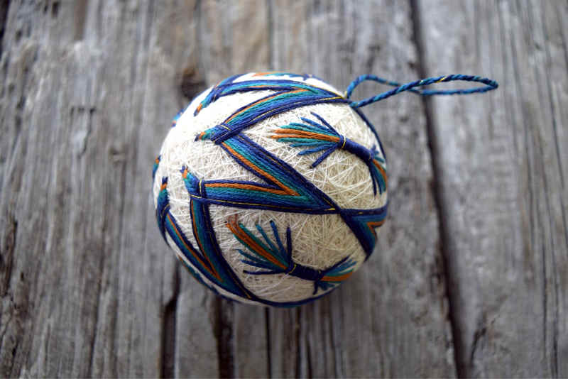 Temari ball 'Peacock' from side, showing blue, teal, and warm yellow-brown design picked out with golden accents against cream colored temari base.