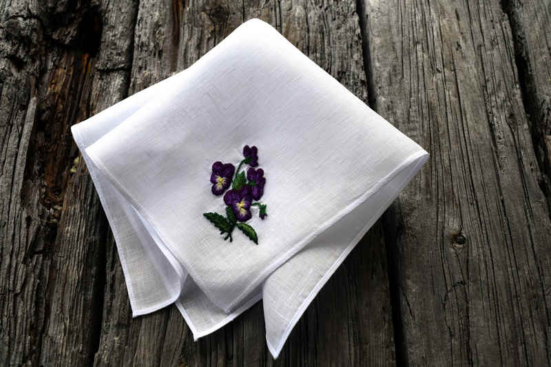 Angled shot of white linen handkerchief, folded, with hand embroidered bunch of johnny jump ups in one corner