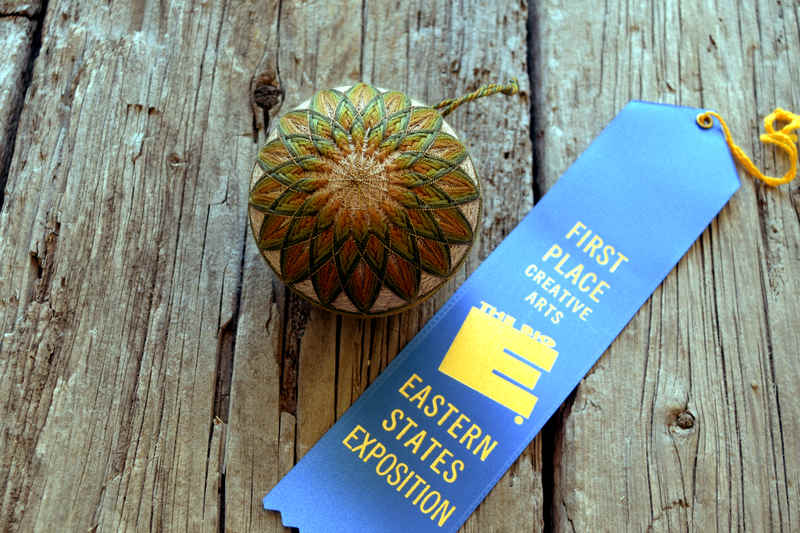 Olive and bronze kiku temari ball 'Summer's End' on wood background next to a blue ribbon from the Eastern States Expo 2022