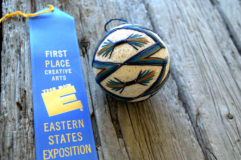 Cream temari ball with blue, teal, and gold design on wood background next to first-place blue ribbon