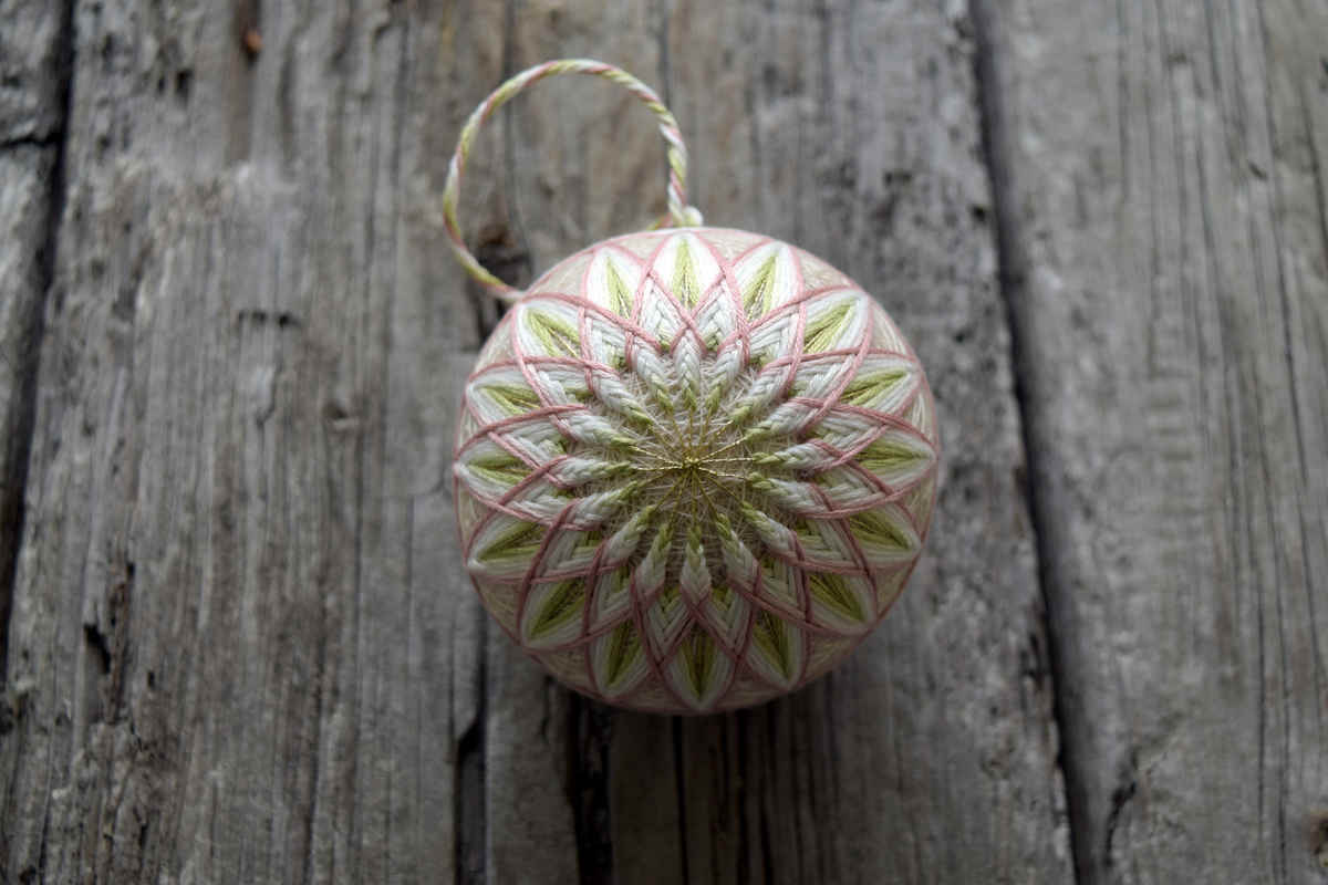 Top down view of hand embroidered temari 'Allium'. Temari is oriented so that the starburst kiku pattern is facing the viewer. Marking lines are gold and design is spring green on the inside shading out to dusty rose. A thread hanger is visible from the top of the ball.