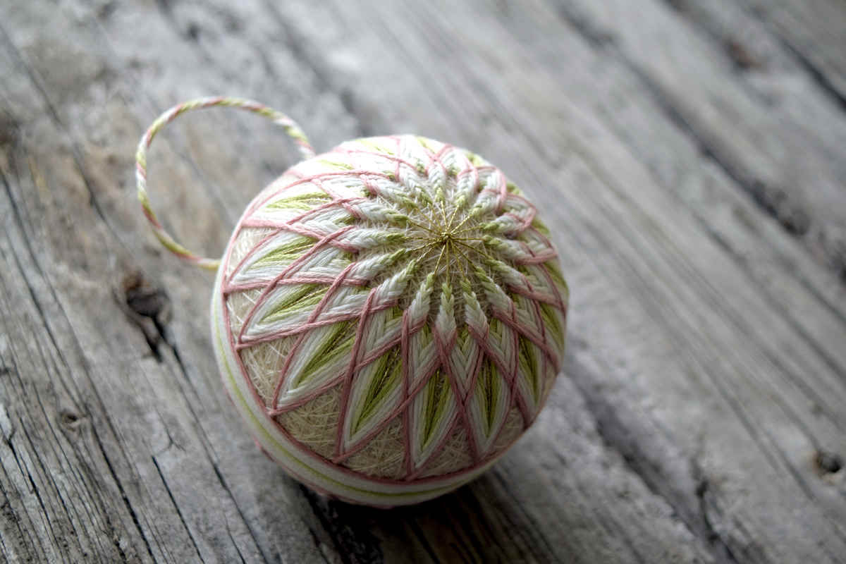 Side view of hand embroidered temari ball 'Allium'. Temari is embroidered in soft shades of cream, spring green, and dusty rose in a starburst pattern. An obi band in the same colors just touches the outer tips of the design