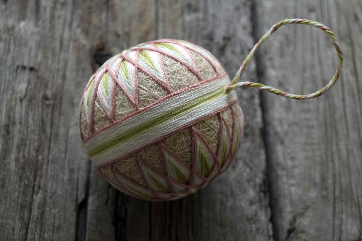 Top-down view of hand embroidered temari ball, centered on obi band and with thread hanger visible as well as points of kiku pattern. Design is worked in pastel shades of dusty rose, spring green, and cream on a sand colored base.