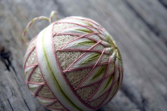 Close up view of an embroidered temari ball. Base of design is sand colored thread, and a starburst pattern is worked on both sides of the sphere with a band of thread in the middle. Embroidery shades from dusty rose through cream to light spring green.