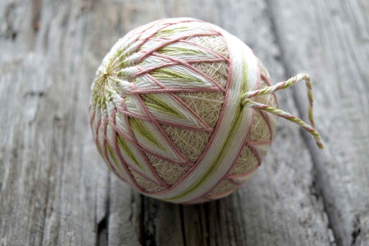 Closeup view of temari ball 'Allium', hand embroidered in soft shades of cream, pink, and green. A thread loop for hanging is attached to the center of the obi band. The poles of the ball are ornamented with many-petaled kiku designs.