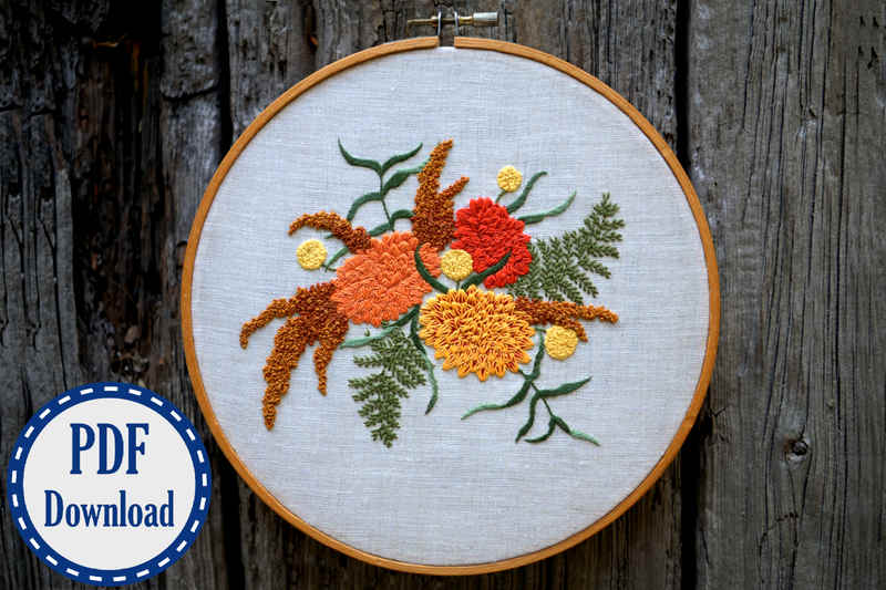 Embroidery hoop with hand stitched bouquet in fall colors. Bouquet has ferns, brown amaranth, and other flowers arranged three colorful chrysanthemums. A badge in the corner says PDF download.