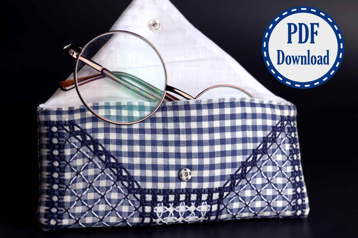 Pair of glasses peeking from a blue and white padded glasses case. Case is made of gingham fabric and is heavily embroidered in chickenscratch embroidery. A badge in the corner says PDF download.