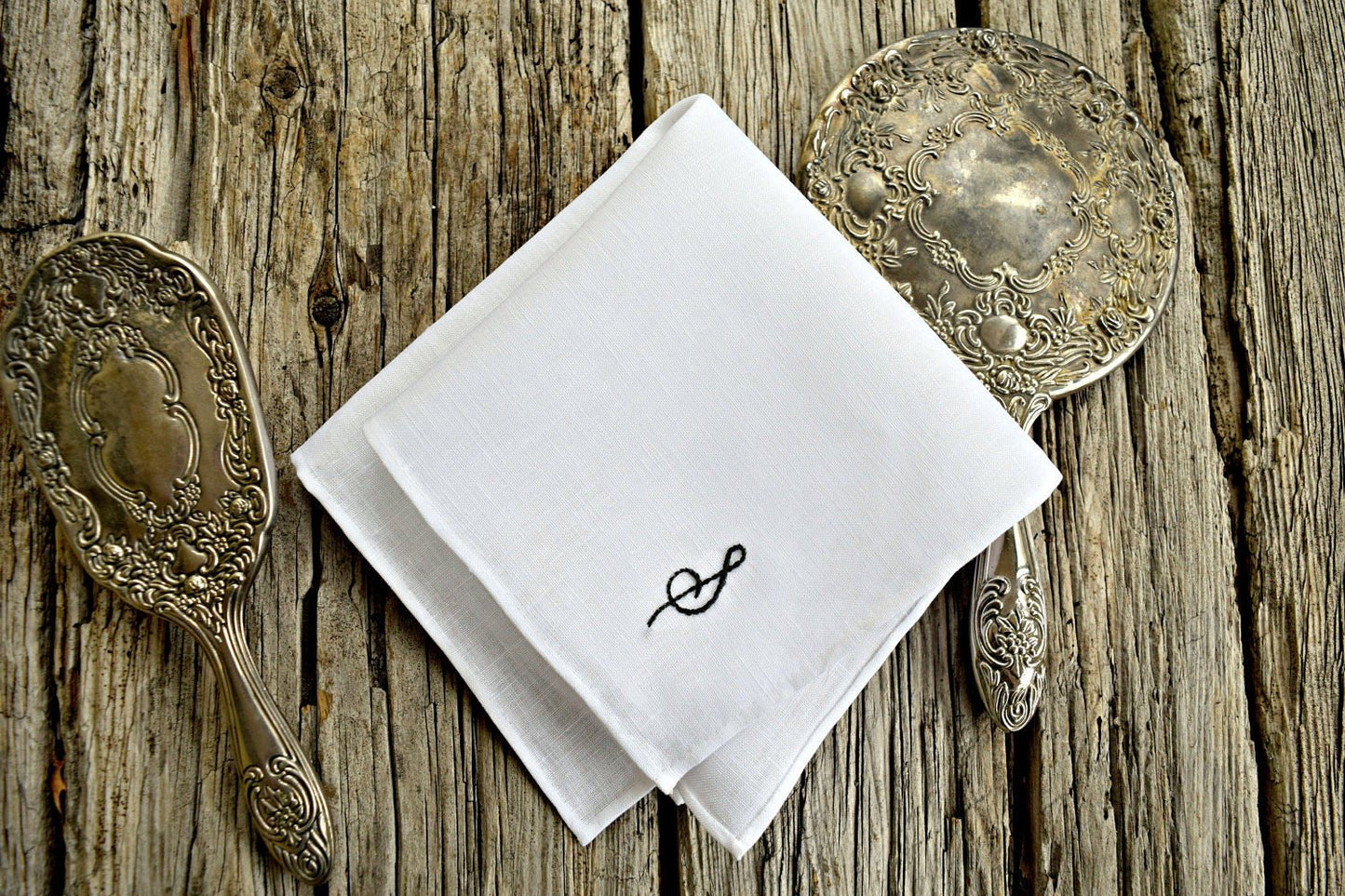 White linen handkerchief with hand embroidered initial between silver mirror and brush