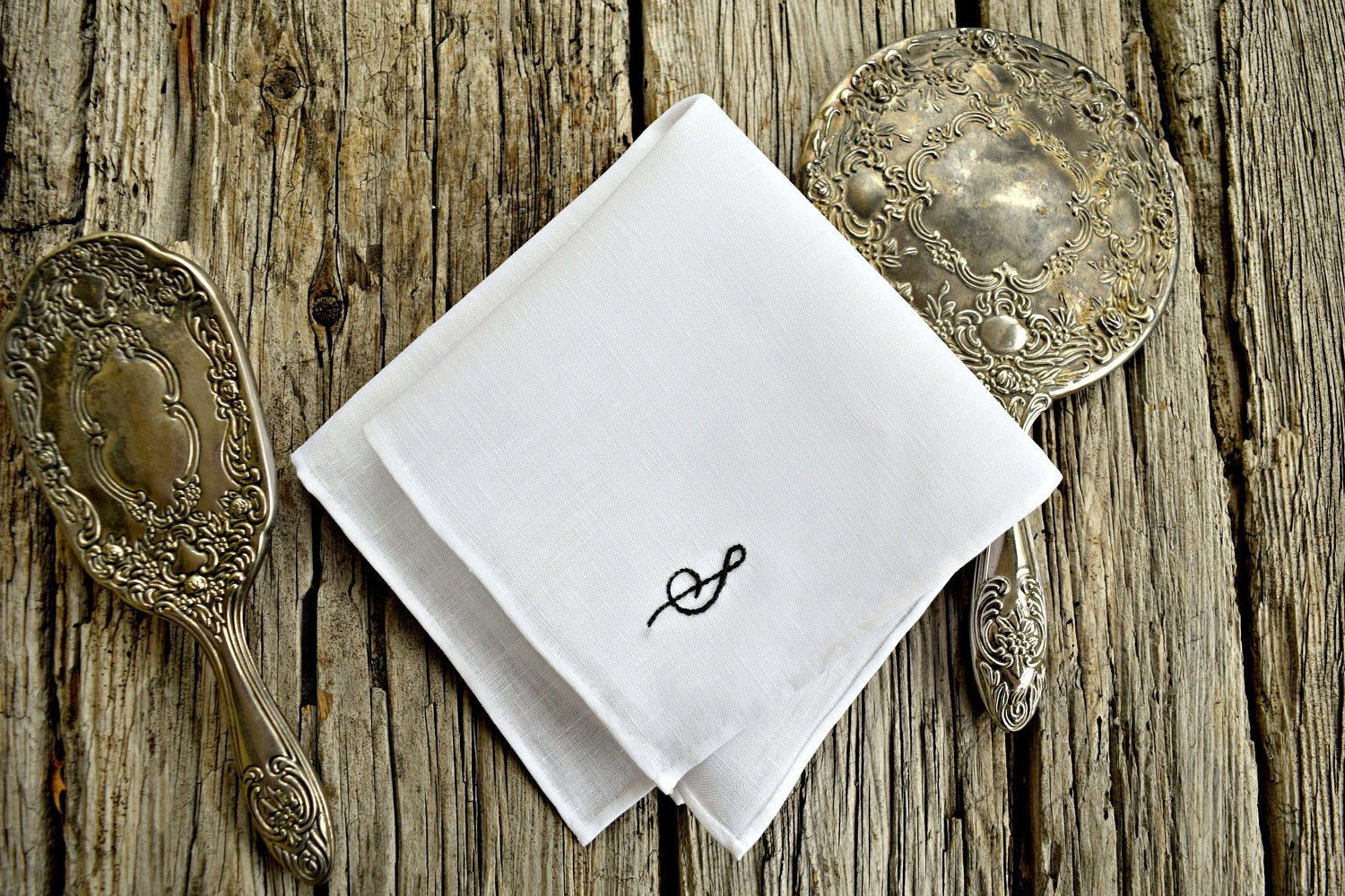 White linen handkerchief with hand embroidered initial between silver mirror and brush