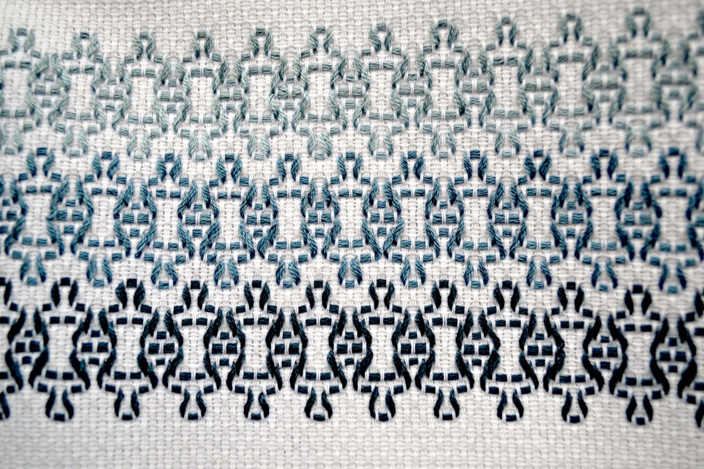 Closeup of repeating motif in huck embroidery done in soft grey blues on white