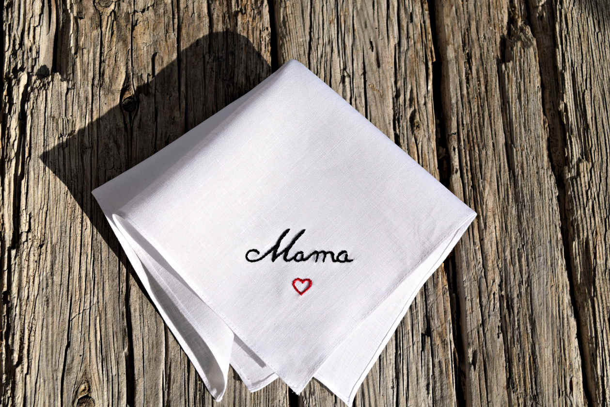 Closeup of linen hankie embroidered in a cursive script. Handkerchief reads 'Mama' in black with a red heart centered below.
