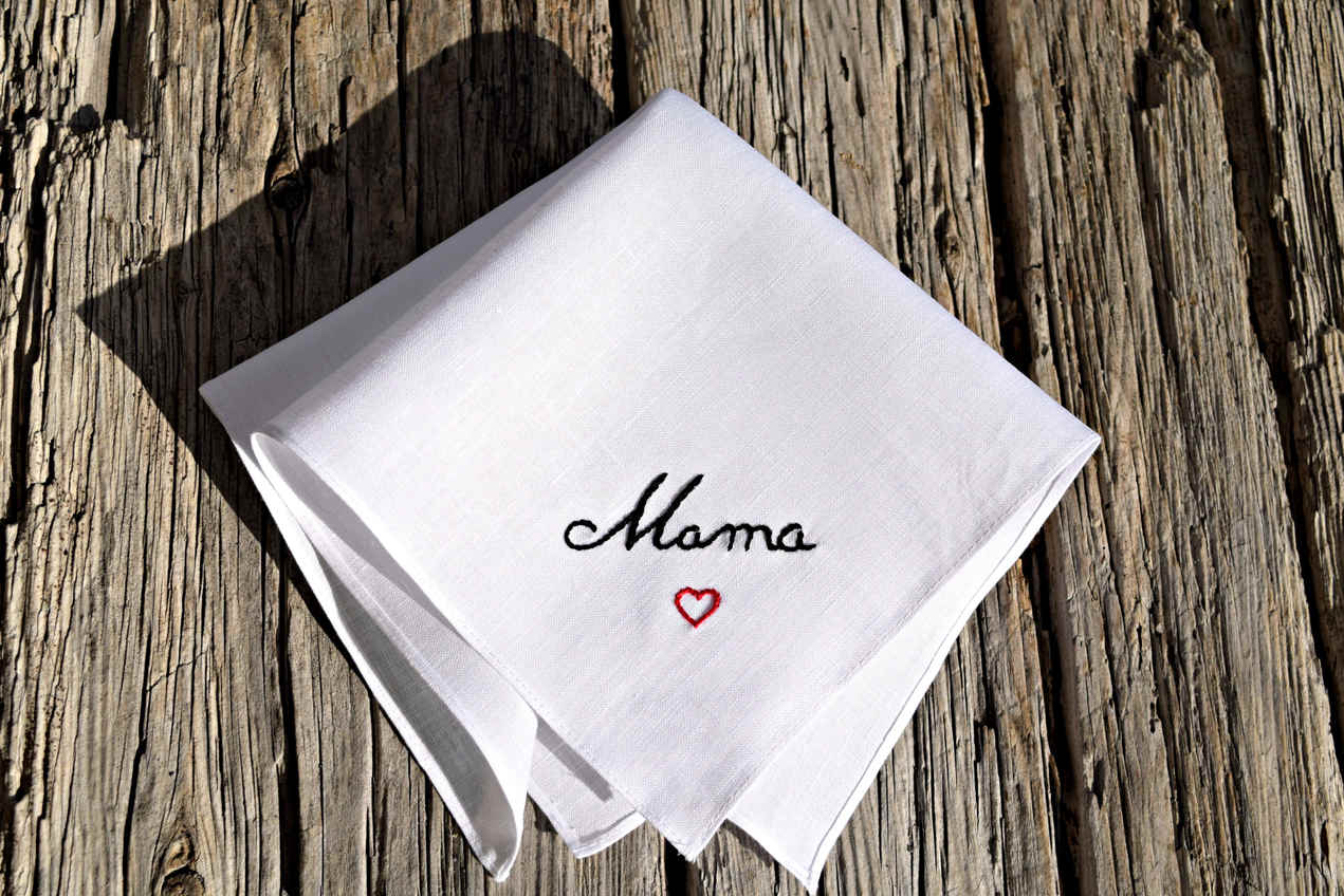 White linen handkerchief folded on wood background, hand embroidered with Mama in black script with red heart below